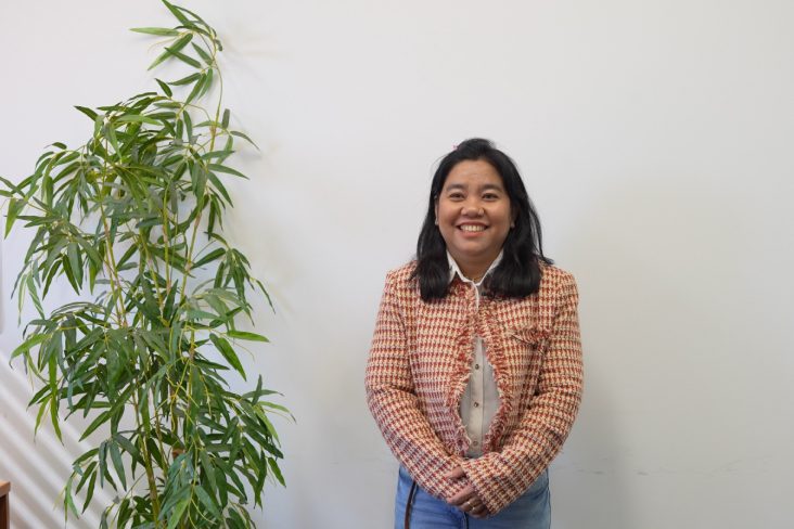 Mariati smiling at the camera with her arms crossed at her waist and a pot plant to the side of her