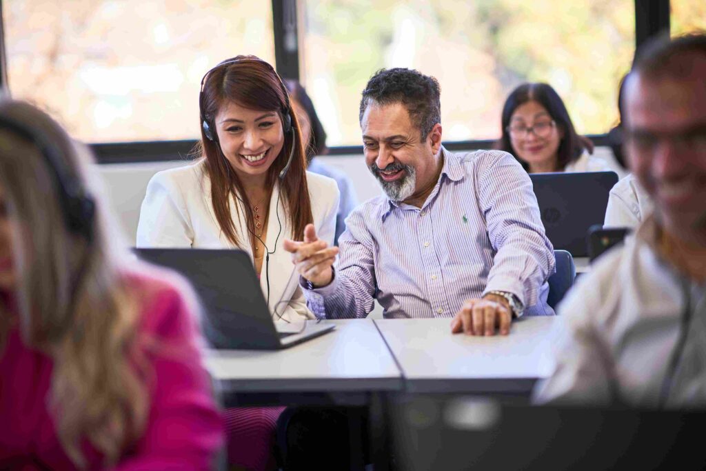 A female student with a headset sitting at a desk next to a teacher pointing at a laptop screen