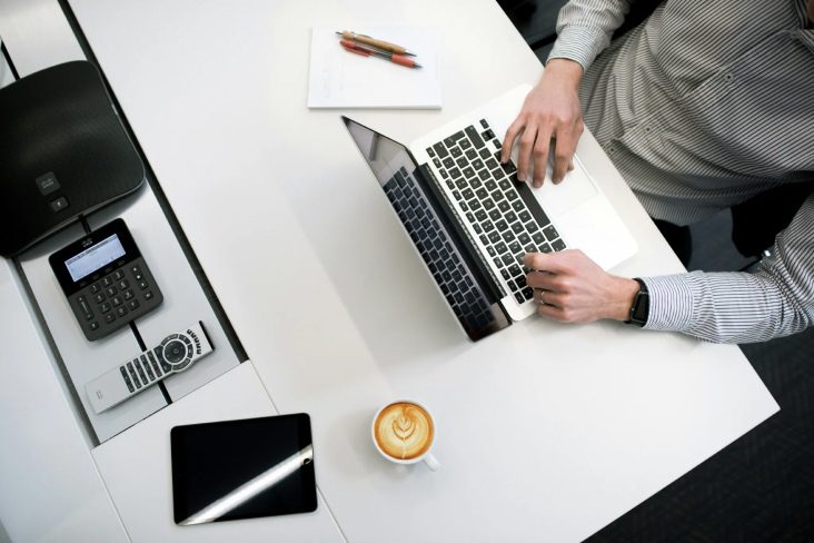 Aerial shot of a person working on a laptop at a desk, with a cup of coffee, paper and pens beside them