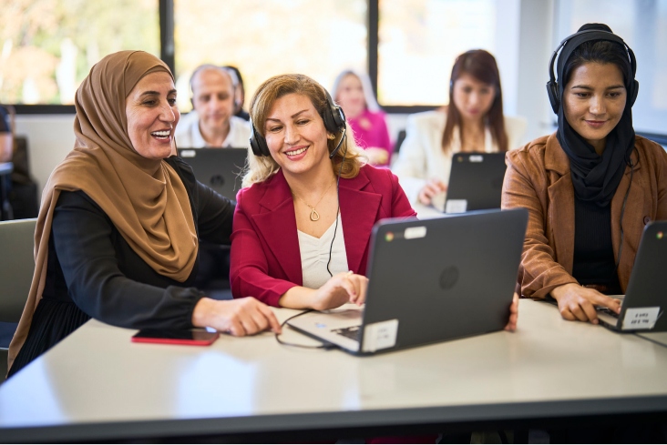 AMEP Student Fozieh sits at a desk with an instructor, using a laptop with headphones over her ears