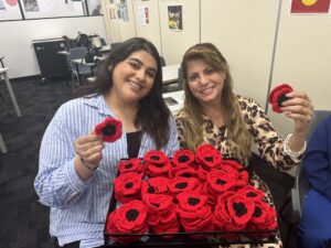 A photo of two women sitting with a container of crocheted poppy wreaths made fro ANZAC day. Both women are holding a poppy in their hand.