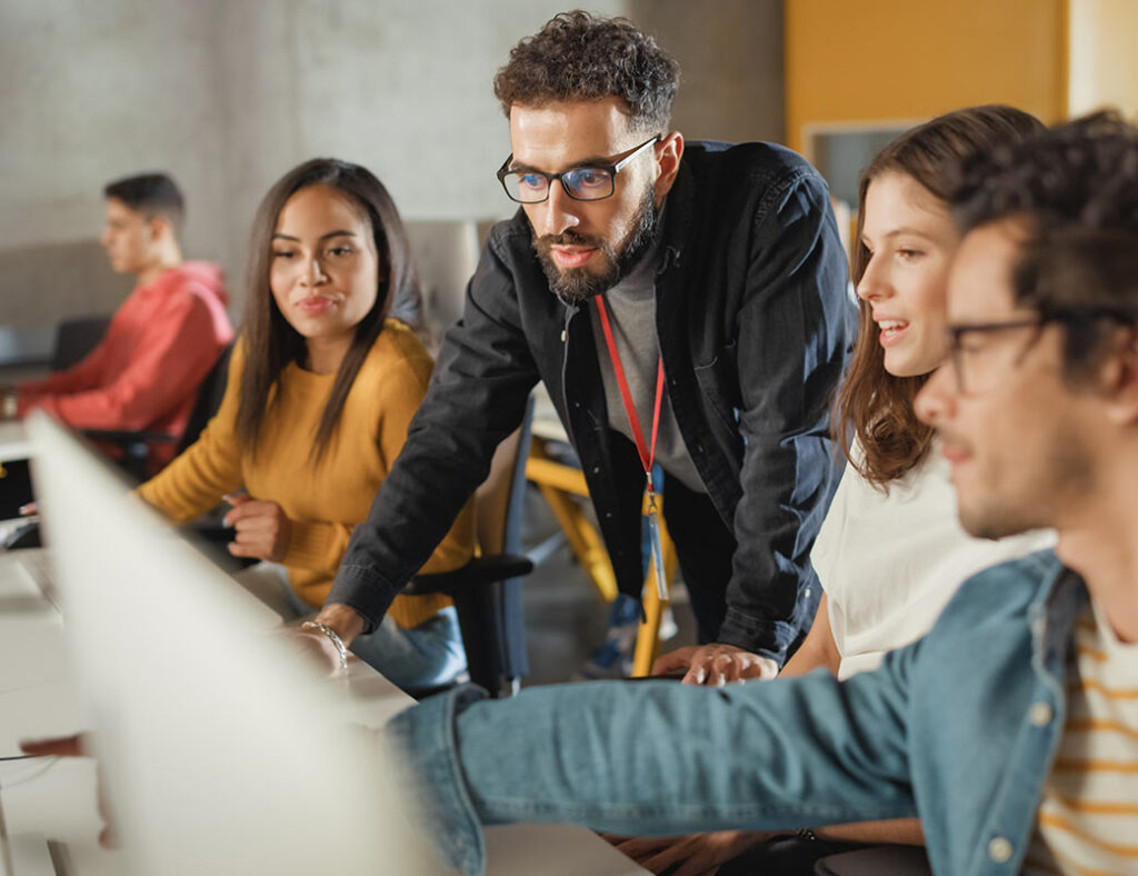 Three students sitting at a long desk and looking towards computer monitors, with a teacher standing over the desk and operating the mouse - Workplace skills (BSB)