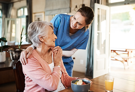 A young female home and aged care worker wearing blue scrubs and standing beside an older woman with her hands placed on her shoulders (440x300)