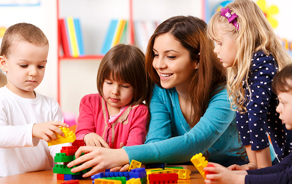 A childcare worker helping four children to build lego blocks at a school desk (600x380)