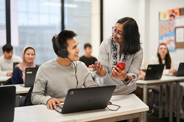 An EAL certificate II teacher holding a smartphone in her hand and talking with a student sitting in front of a laptop and wearing over ear headphones (600x400)