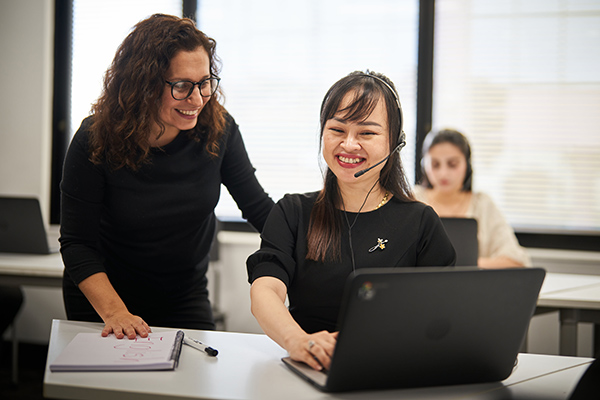 An EAL certificate III teacher standing beside a student sitting in front of a laptop and wearing a customer service headset (1200x800)