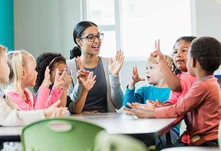 Six children sitting at a table with a female childcare worker and holding their hands up in the air (440x300)