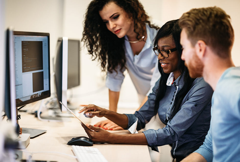 Two NSF students sitting at a desk in front of a desktop monitor and looking towards a tablet screen, with a teacher standing over them and guiding them (980x660)
