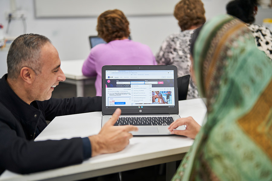 An NSF teacher kneeling at a desk and helping an NSF Computer & Digital Skills short course student (900x600).