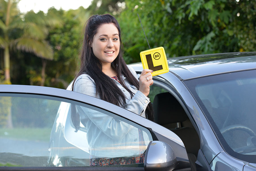 A young female student standing beside the driver-side door of a car and holding a learner's plate in her hand (900x600).