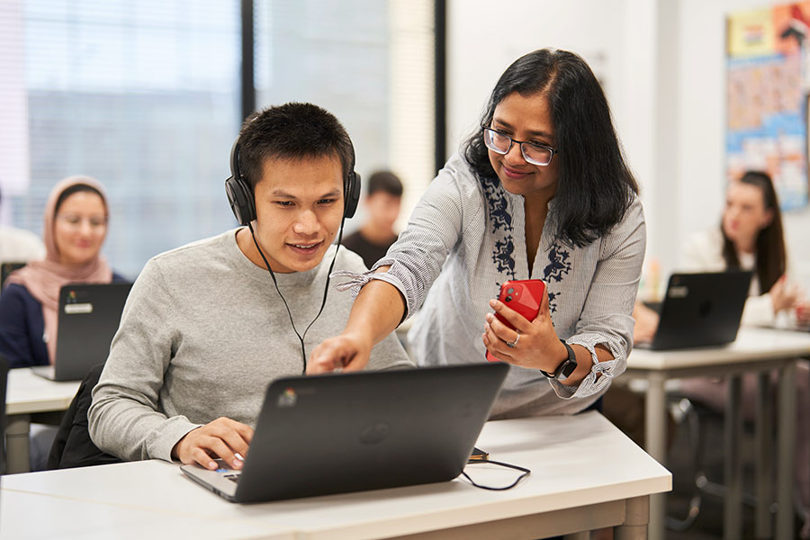 A student sitting at a desk in front of a laptop and wearing over-ear headphones. A Navitas Skilled Futures teacher is standing beside the desk and pointing towards the laptop screen (900x600).