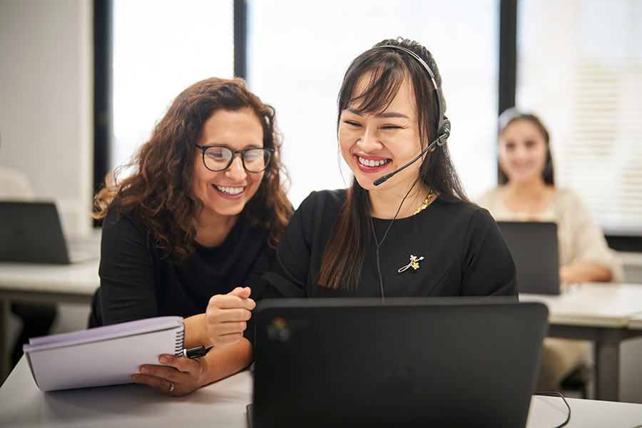A young NSF student sitting in front of a laptop and wearing a customer service headset while her teacher smiles and looks towards her laptop screen (900x600).