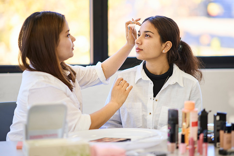 Two female beauty service students sitting together. One student is applying makeup to the forehead of the other student (900x600).