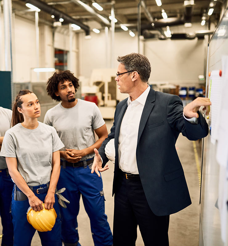 A man in a business suit pointing towards a whiteboard while factory workers wearing blue pants and hard hats observe (740x800).