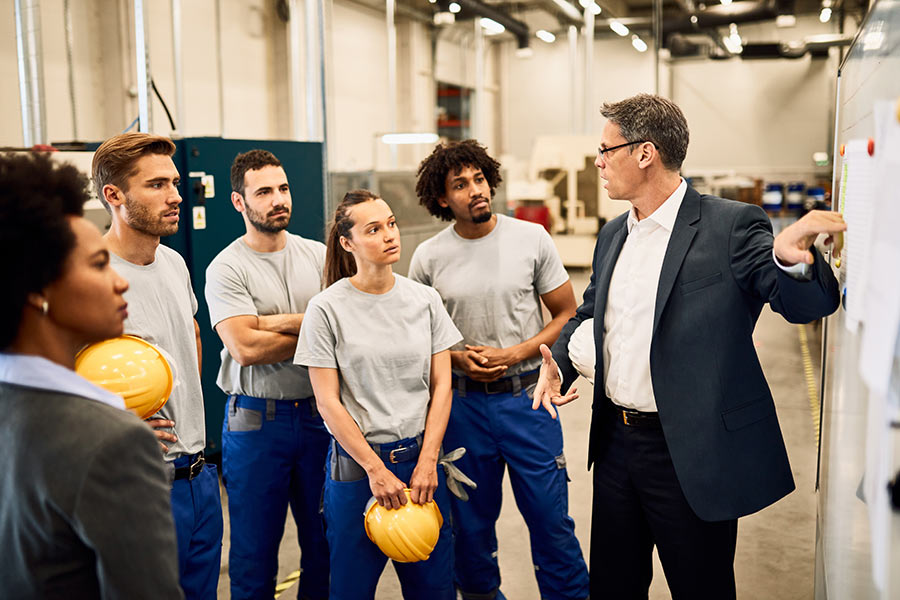 A man in a business suit pointing towards a whiteboard while factory workers wearing blue pants and hard hats observe (900x600).
