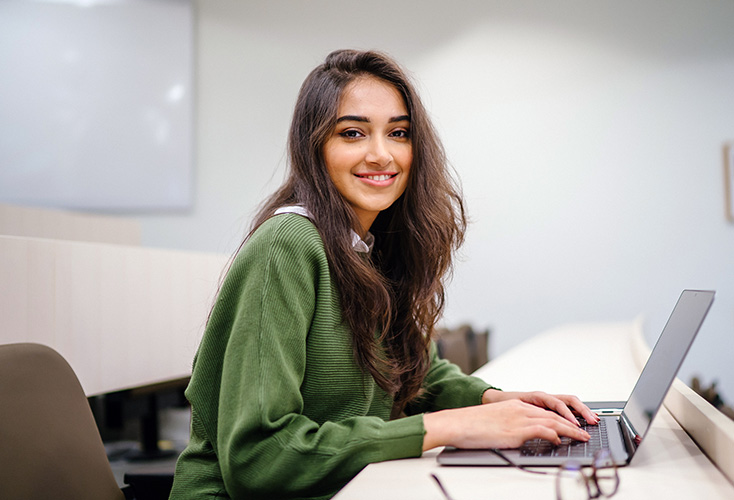 A young female NSF student sitting at a desk in a classroom, looking at a laptop screen and wearing a customer service headset (440x300).