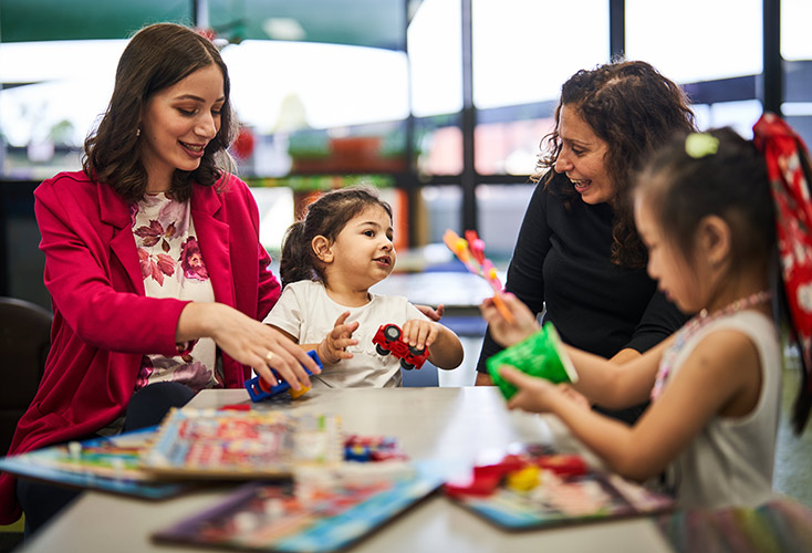 Two female childcare workers sitting at a table with two children as they play with toys (734x500).