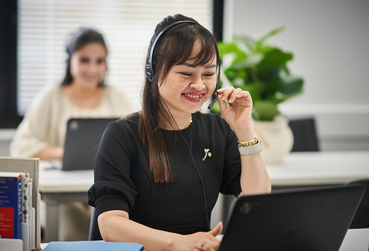 A young female NSF student sitting at a desk in a classroom, looking at a laptop screen and wearing a customer service headset (734x500).