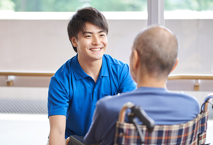 A young male care industries student kneeling in front of a man who is sitting in a wheelchair (734x500).