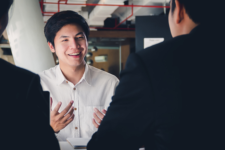 A young male workplace communication student sitting at a table across from two office workers wearing suits (900x600).
