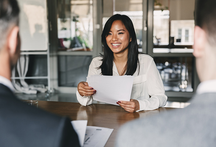 A young woman sitting in a job interview with papers in her hands, sitting across a table from two people wearing business attire (734x500).
