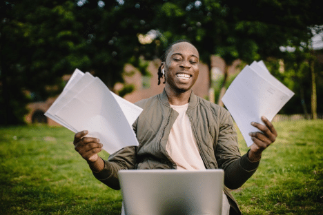 A happy looking man sitting in front of a laptop with papers in each hand