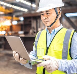Man wearing Hi-Vi vest and hard hat, reviewing a laptop at a construction site.