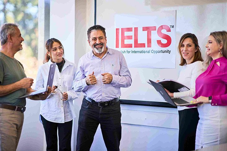 A teacher from the IELTS preparatory course standing in front of a whiteboard, with several students standing around him and holding laptops in their hands (732x488).