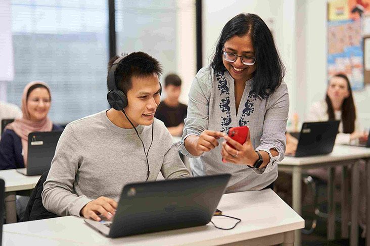 A male student wearing a headset and sitting at a desk in front of a laptop. A teacher is showing something to the student on the screen of her smartphone (732x488).