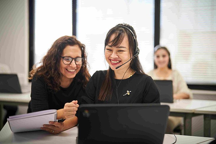 A SEE Program teacher standing beside a student sitting in front of a laptop and wearing a customer service headset (732x488)