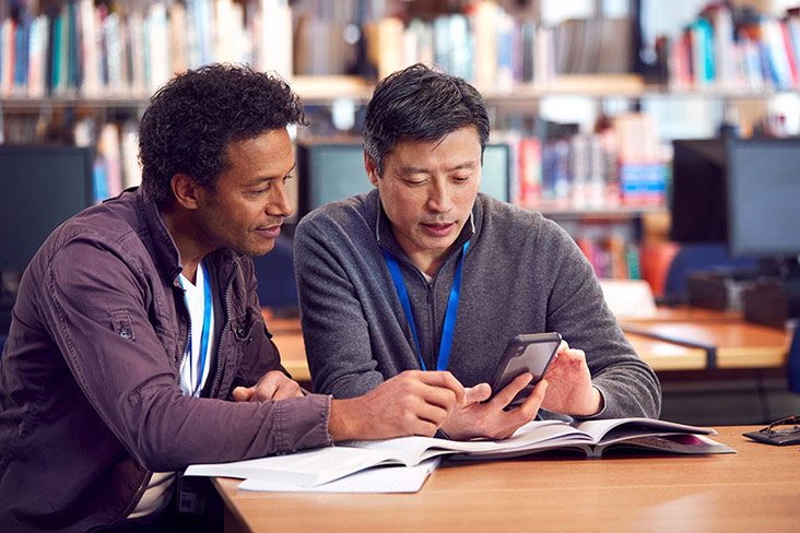 A student sitting at a desk with a SEE student support officer and looking towards a mobile phone in the support officer's hands (732x488).