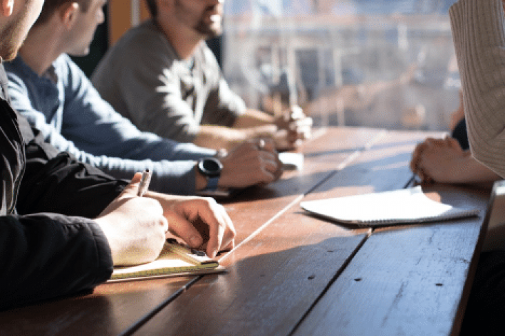 A close up photograph of studnets taking notes on a notepad on a wooden desk with the sun setting in the background