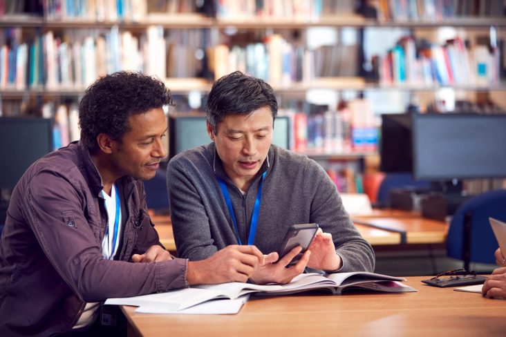Teacher With Mature Male Adult Student Using Mobile Phone At Table Working In College Library